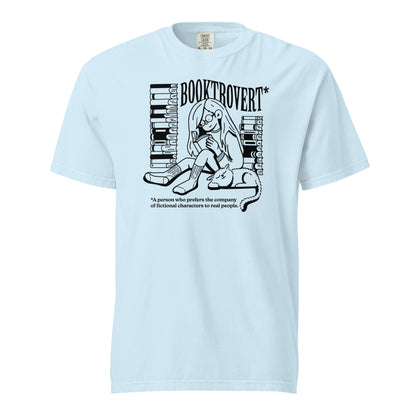 Chambray Blue Comfort Colors t-shirt with the text "Booktrovert." A woman holds a book reading, with her cat sitting next to her sleeping. Behind her are two piles of books. The bottom reads "A person who prefers the company of fictional characters to read people."