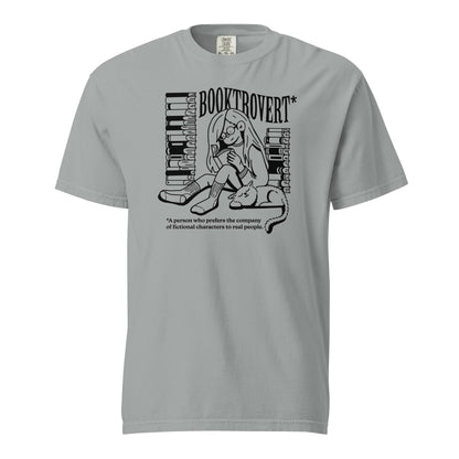 Granite Gray Comfort Colors t-shirt with the text "Booktrovert." A woman holds a book reading, with her cat sitting next to her sleeping. Behind her are two piles of books. The bottom reads "A person who prefers the company of fictional characters to read people."