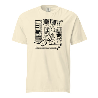 Ivory White Comfort Colors t-shirt with the text "Booktrovert." A woman holds a book reading, with her cat sitting next to her sleeping. Behind her are two piles of books. The bottom reads "A person who prefers the company of fictional characters to read people."