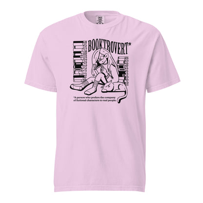 Orchid Light Purple Pink Comfort Colors t-shirt with the text "Booktrovert." A woman holds a book reading, with her cat sitting next to her sleeping. Behind her are two piles of books. The bottom reads "A person who prefers the company of fictional characters to read people."