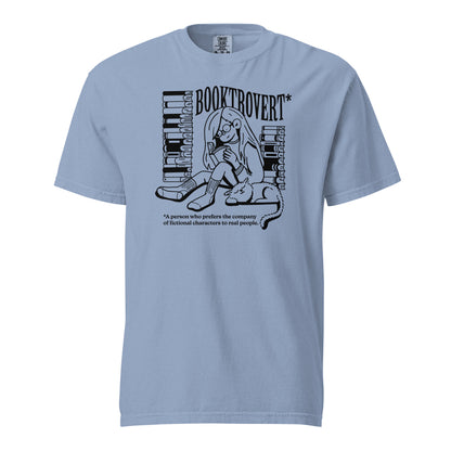 Washed Denim Blue Comfort Colors t-shirt with the text "Booktrovert." A woman holds a book reading, with her cat sitting next to her sleeping. Behind her are two piles of books. The bottom reads "A person who prefers the company of fictional characters to read people."