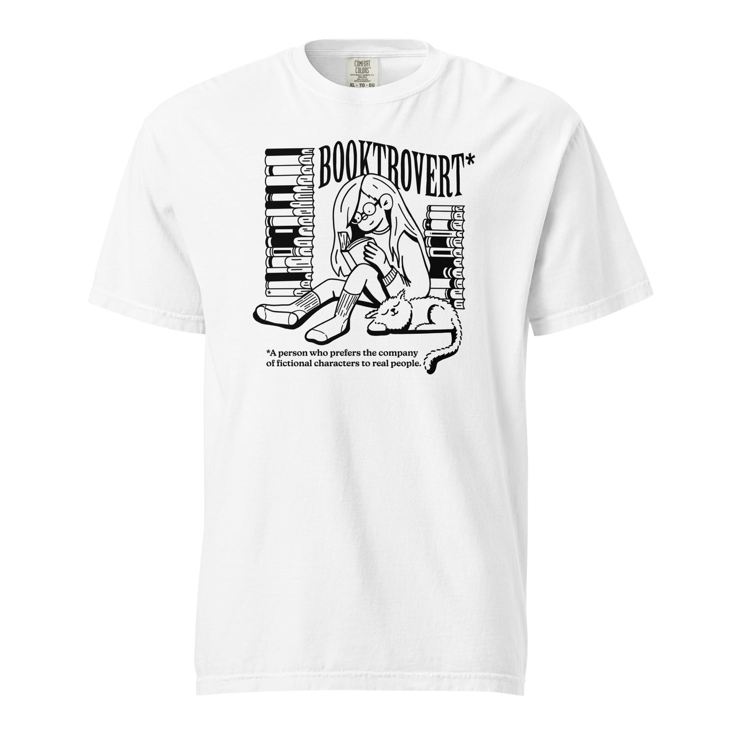 White Comfort Colors t-shirt with the text "Booktrovert." A woman holds a book reading, with her cat sitting next to her sleeping. Behind her are two piles of books. The bottom reads "A person who prefers the company of fictional characters to read people."