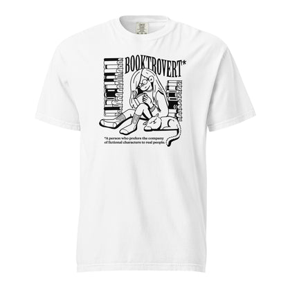 White Comfort Colors t-shirt with the text "Booktrovert." A woman holds a book reading, with her cat sitting next to her sleeping. Behind her are two piles of books. The bottom reads "A person who prefers the company of fictional characters to read people."